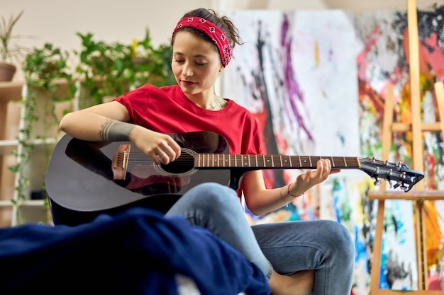 Entspannte junge Frau, die Gitarre spielt, wie man in Freizeitkleidung auf dem Bett sitzt