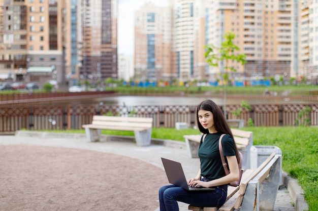 Entspannte junge Frau in einem grünen T-Shirt und mit einem Rucksack, der auf einer Holzbank sitzt, Kaffee trinkt und auf ihrem Laptop stöbert