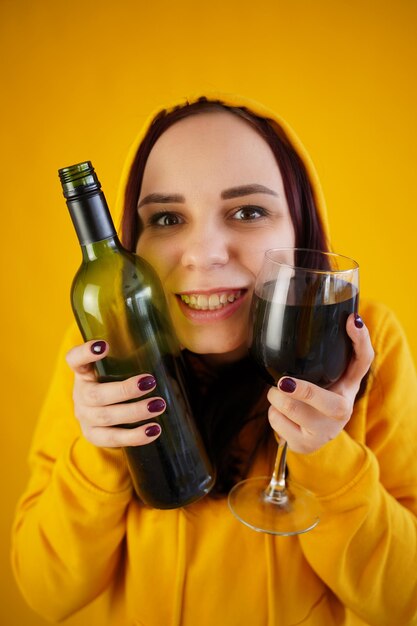 Entspannte junge Frau posiert mit Flasche und Glas Rotwein auf gelbem Hintergrund Erwachsene glückliche Brünette drückt Alkohol ins Gesicht