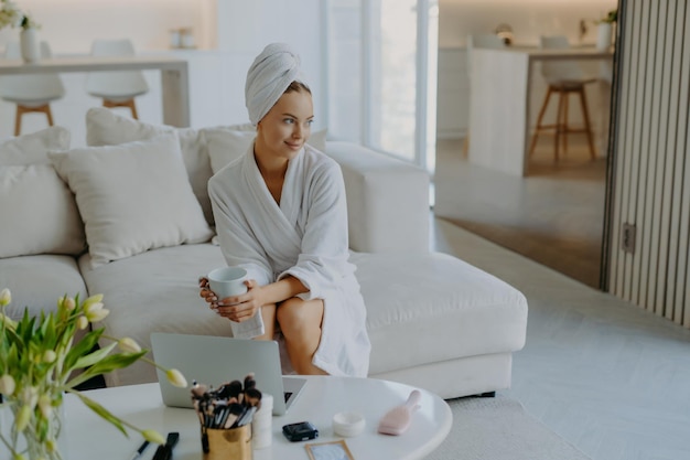 Entspannte, nachdenkliche Frau im Bademantel und eingewickeltem Handtuch auf dem Kopf sitzt auf dem Sofa mit einer Tasse Getränk in der Nähe des Tisches mit Kosmetikprodukten und sieht nachdenklich beiseite, posiert vor dem gemütlichen Wohninterieur