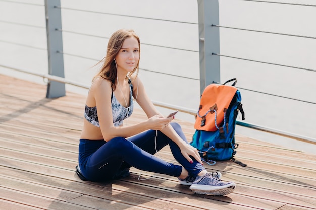 Entspannte reizende Frau mit dem dunklen Haar, gesunde reine Haut, gekleidet in der Sportkleidung, hält Handy für Kommunikation