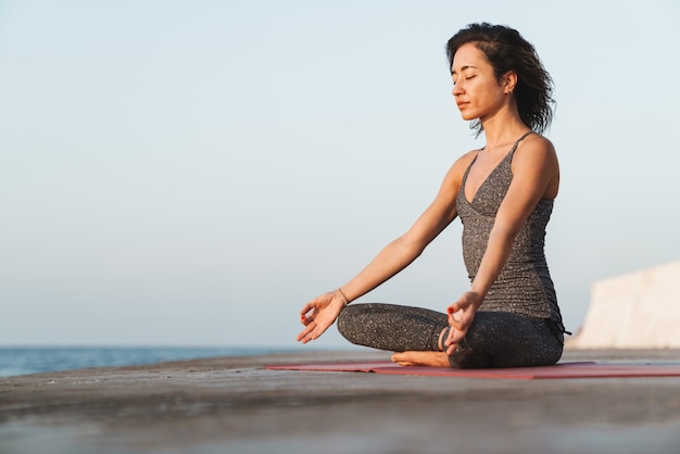 Entspannte Sportlerin im Trainingsanzug, die in Lotuspose auf dem Pier sitzt und morgens am Meer Yoga praktiziert