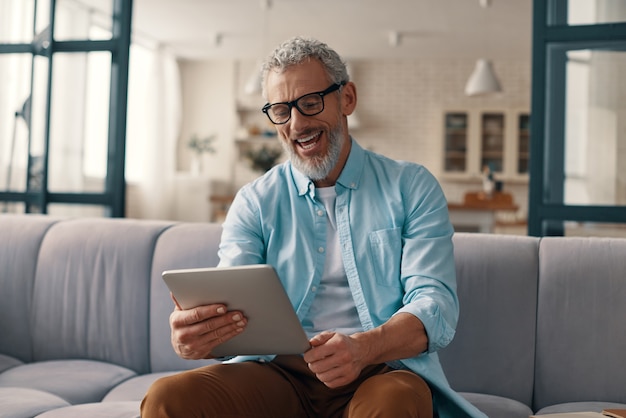 Entspannter älterer Mann, der ein digitales Tablet verwendet und lächelt, während er zu Hause auf dem Sofa sitzt