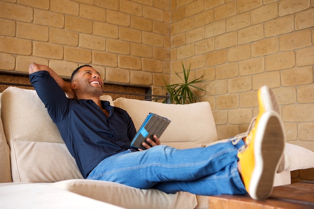 Entspannter junger schwarzer Mann, der im Wohnzimmer mit digitaler Tablette sitzt