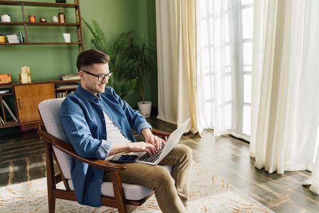 Entspannter Mann in Jeanshemd arbeitet an einem Laptop in einem gemütlichen, sonnigen Raum