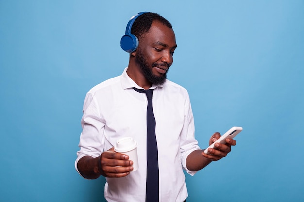 Entspannter Mann mit drahtlosen Kopfhörern, der soziale Medien auf dem Smartphone durchsucht, während er eine Tasse Kaffee zum Mitnehmen hält. Geschäftsmann in einer Pause Musikhören über das Ohr-Headset mit Blick auf das Smartphone.