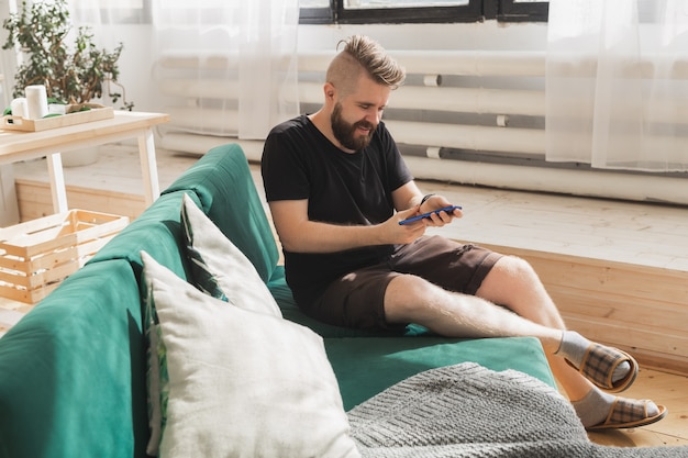 Entspannter Mann mit einem Smartphone, der zu Hause auf der grünen Couch im Wohnzimmer sitzt