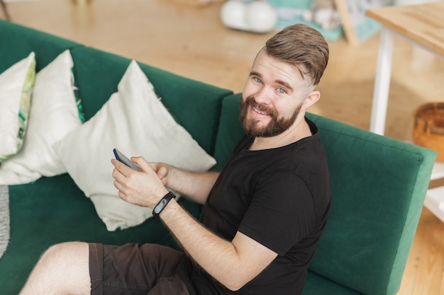 Entspannter Mann mit einem Smartphone, der zu Hause auf der grünen Couch im Wohnzimmer sitzt