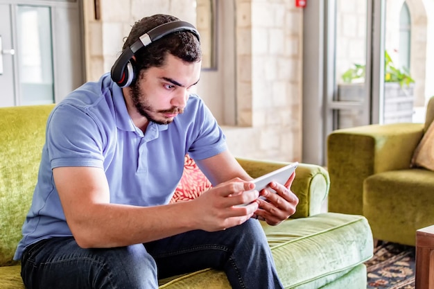 Entspannter Mann mit Kopfhörern, der sich Videos auf dem Tablet ansieht, das zu Hause auf einer Couch im Wohnzimmer sitzt