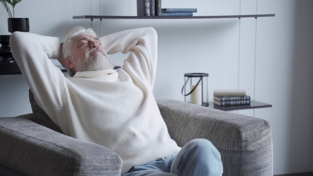 Entspannter reifer Mann mit einem grauen Bart zu Hause, der auf einem Stuhl sitzt
