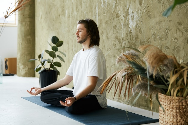 Entspannter ruhiger Mann, der am modernen Designhaus sitzt und meditiert, reizender Mann in Yoga-Pose und Konzentration