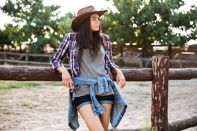 Entspanntes hübsches cowgirl der jungen frau mit hut, das auf ranch steht und ruht