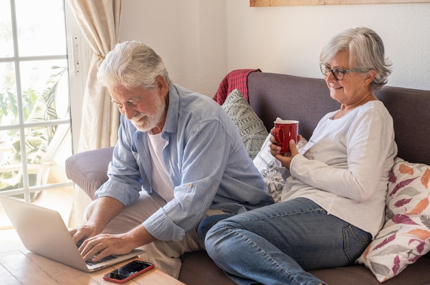Entspanntes kaukasisches älteres Paar, das zu Hause auf Sofa unter Verwendung des Laptops sitzt Zwei ältere Rentner, die Technologie und Soziales genießen