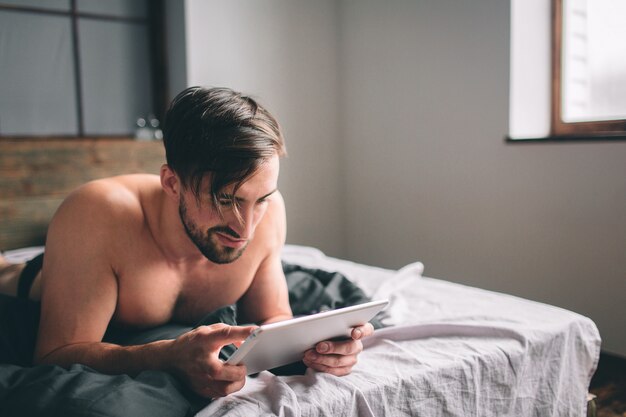 Entspannung des jungen Mannes auf dem Bett und unter Verwendung der digitalen Tablette