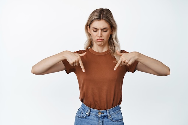 Enttäuschte blonde Frau, die mit dem Finger zeigt und die Stirn runzelt, weil sie eine traurige Reaktion auf etw schlechten weißen Hintergrund nicht mag