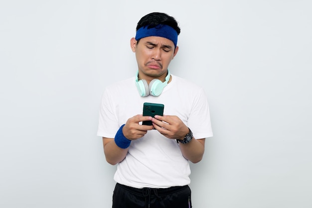 Enttäuschter junger asiatischer sportlicher Mann in blauem Stirnband und weißem T-Shirt mit Kopfhörern mit Handy und Lesen schlechter Nachrichten isoliert auf weißem Hintergrund Trainingssportkonzept