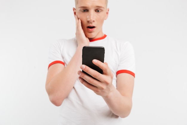 enttäuschter Teenager in Freizeitkleidung, die das Gesicht berührt und den Schock ausdrückt, während er das auf Weiß isolierte Smartphone verwendet