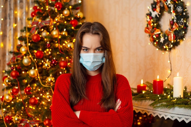 Enttäuschung junge Frau in medizinischer Maske, die mit gefalteten Händen vor dem Weihnachtsbaum steht Konzept von Krankheiten und Viren in den Ferien