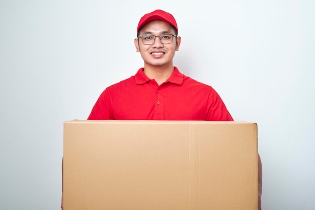 Entusiasmado jovem correio asiático em uniforme vermelho e boné parece surpreso segurando a caixa de pedido do cliente