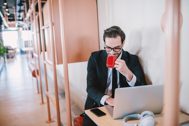 Entusiasta apuesto empresario barbudo caucásico en traje y con anteojos sentado en la mesa, tomando café y terminando el informe. Interior de la empresa.