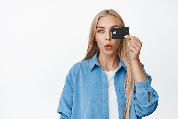 Entusiasta chica rubia con tarjeta de crédito en el ojo, con curiosidad, de pie en traje casual en blanco.