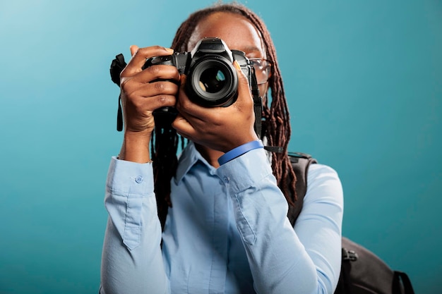 Entusiasta de fotografia de adulto jovem afro-americano com dispositivo de fotografia moderno tirando fotos em fundo azul. Fotógrafo confiante e criativo tirando fotos enquanto aproveita o tempo de lazer.
