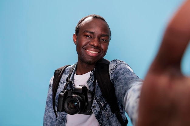 Entusiasta de fotografia feliz com dispositivo de foto profissional e tirando selfie em fundo azul. fotógrafo confiante com câmera dslr e mochila curtindo férias.