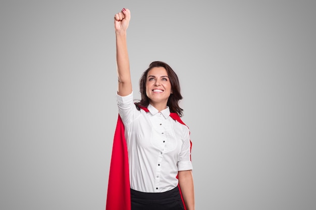 Entusiasta empresaria adulta con camisa blanca y capa roja de superhéroe levantando la mano con el puño cerrado mientras celebra el logro contra el fondo gris