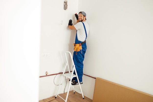 Entusiasta manitas electricista sonriendo a la cámara mientras trabaja en la instalación de enchufe en la pared