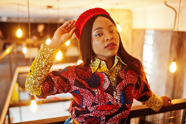 Entusiasta mujer afroamericana en traje de moda color con boina roja escalofriante en acogedor café.