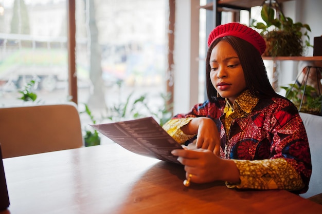 Entusiasta mujer afroamericana vestida de moda con boina roja relajándose en un acogedor menú de lectura de café