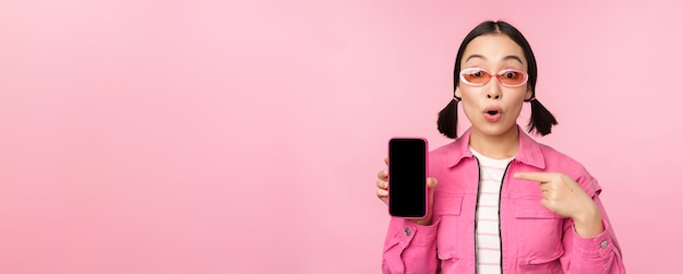 Entusiasta mujer asiática con ropa elegante gafas de sol apuntando con el dedo a la pantalla del teléfono móvil que muestra la aplicación del teléfono inteligente de pie sobre fondo rosa