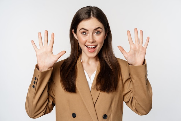 Entusiasta mujer de negocios con traje, levantando las manos con las palmas hacia arriba, número diez docenas de gestos y sonriendo, de pie sobre fondo blanco.