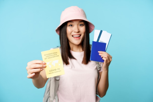 Entusiasta turista asiático, niña muestra certificado de vacunación internacional covid-19, pasaporte con dos boletos, viaje después de la vacuna contra el coronavirus, fondo azul