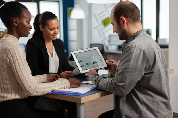 Entusiastas empresarios diversos leyendo el informe financiero anual sentado a la mesa en la moderna oficina de negocios de inicio sosteniendo la tableta y sonriendo