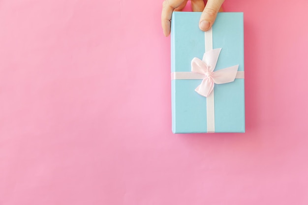 Entwerfen Sie einfach die Hand einer weiblichen Frau, die eine blaue Geschenkbox isoliert auf rosa pastellfarbenem, trendigem Hintergrund hält ...