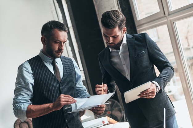 Entwicklung eines neuen Projekts. Zwei junge moderne Männer in formeller Kleidung arbeiten zusammen, während sie drinnen stehen standing