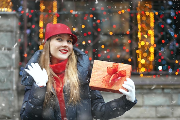 Entzückende blonde Frau mit einer Geschenkbox auf dem Straßenweihnachtsmarkt während des Schneefalls. Freiraum