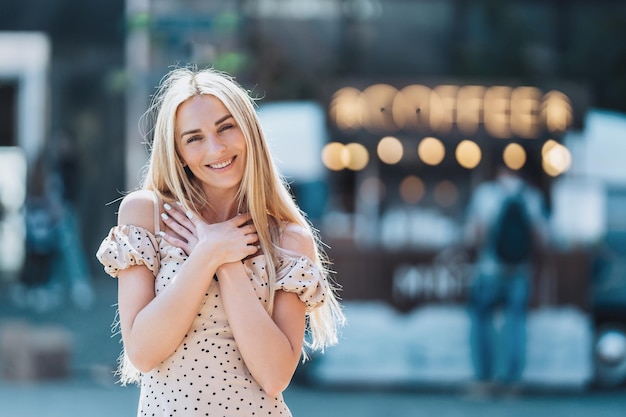 Entzückende blonde kaukasische Frau mit langen, lockeren Haaren, die mit gefalteten Händen auf der Brust breit lächelt, blickt auf die Kamera vor verschwommenem City Food Court im Hintergrund Zufriedenes schwedisches Mädchen im Sommer