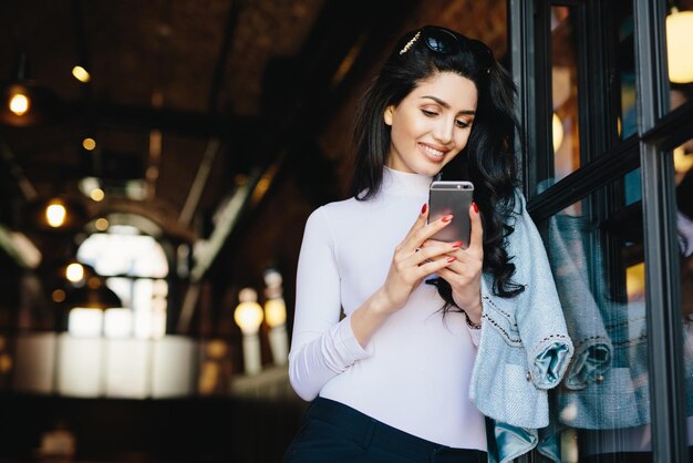 Entzückende brünette Frau mit fröhlichem Aussehen, die eine Sonnenbrille auf dem Kopf hat und eine weiße Bluse trägt, die über einem modernen Café-Interieur sitzt und mit dem Handy den Newsfeed auf ihren Konten in sozialen Netzwerken überprüft