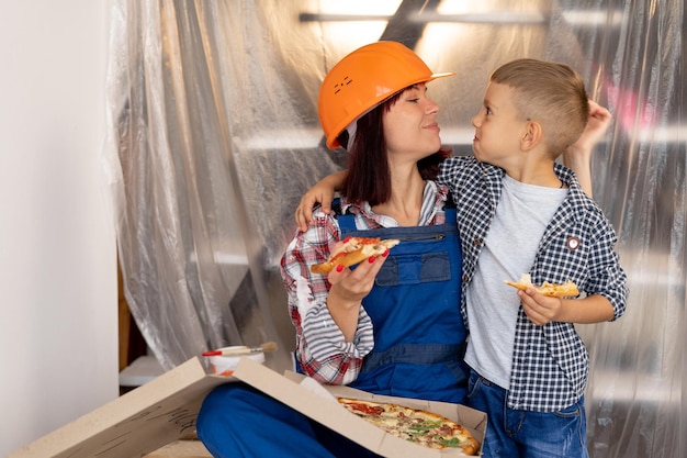 Entzückende Familienmutter und Sohn ruhen sich aus und essen köstliche italienische Pizza, müde nach der Renovierung in einem neuen Haus. Eine Frau mit Schutzhelm und ein Junge schauen mit Liebe nach Reparatur- und Handwerkerkonzept