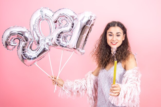 Entzückende festlich gekleidete brünette Frau mit dem lockigen Haar betrachtet eine ausgelöschte Feuerwerkskerze mit silbernen Luftballons für das Neujahrskonzept