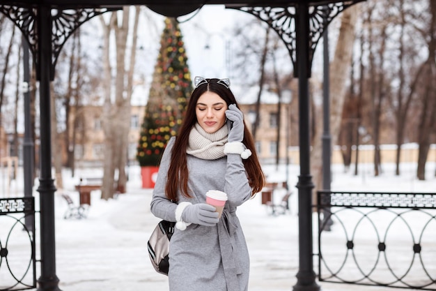 Entzückende Frau spricht auf dem Smartphone im Freien im Winterpark vor dem Weihnachtsbaum, Frau hält eine Kaffeetasse in der Hand.