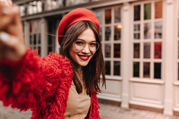 Entzückende gebräunte Dame mit Brille in roter Wolljacke, beigefarbenem Oberteil und heller Baskenmütze macht Selfie auf der Straße