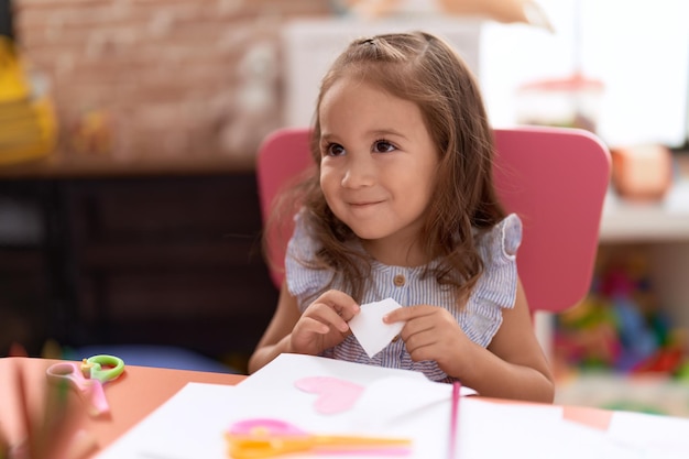 Entzückende hispanische Studentin, die im Kindergarten auf dem Tisch sitzt und Papier schneidet