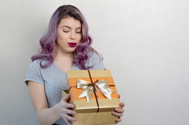 Entzückende junge Frau mit lila Haaren und hellem Make-up mit Geschenkbox, Studioaufnahme auf grauem Hintergrund. Platz für Text