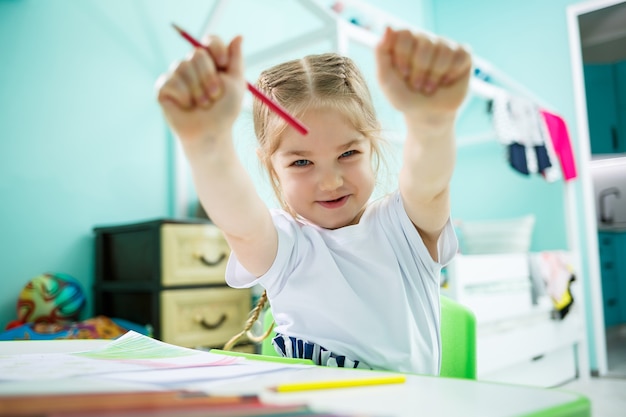 Entzückende Kleinkindmädchenzeichnung mit Bleistiften zu Hause am Tisch sitzend. Kreatives Kind, das in einem Raum sitzt und das Zeichnen lernt. Kleinkindmädchen, das zu Hause Hausaufgaben macht.