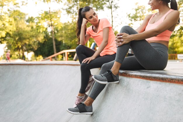 entzückende schöne Frauen in der Sportbekleidung, die mit Wasserflaschen auf Betonsportplatz sitzen und sprechen