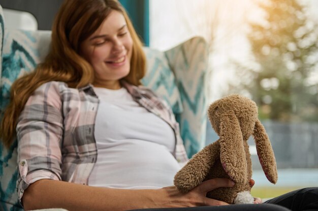 Entzückende schwangere Frau sitzt mit einem Plüschkaninchen und träumt von einem zukünftigen Kind zu Hause in der Nähe des Fensters mit Blick auf den Garten