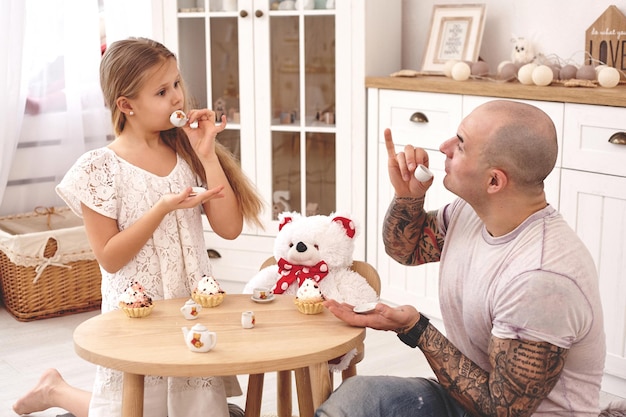 Entzückende Tochter, die ein weißes Kleid trägt, mit ihrem liebevollen Vater trinken sie Tee aus einem Spielzeugk...